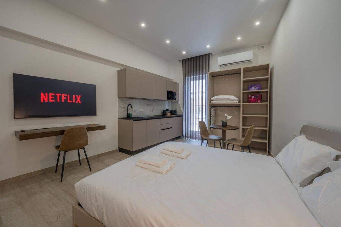 Ein modernes Hotelzimmer mit einem großen Bett, einem Schreibtisch mit Stuhl, einer Küchenzeile, einem an der Wand befestigten Fernseher und einem Kleiderschrank.