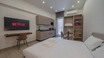 Una habitación de hotel moderna con una cama grande, un escritorio con una silla, una kitchenette, un televisor montado en la pared y un armario.