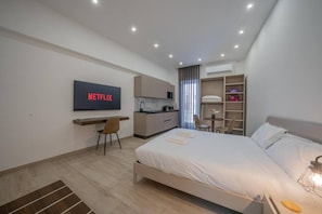 Quarto de hotel moderno com cama, escrivaninha, TV exibindo 'NETFLIX', cozinha compacta e closet espaçoso.