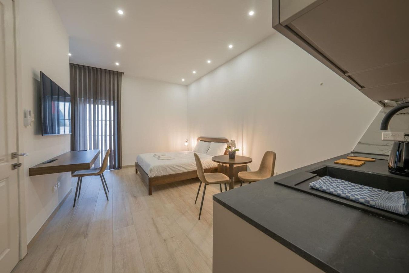 A compact hotel room with a bed, a desk, a chair, a small table, and a kitchenette area.