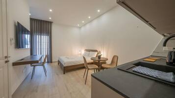 A compact hotel room with a bed, a desk, a chair, a small table, and a kitchenette area.