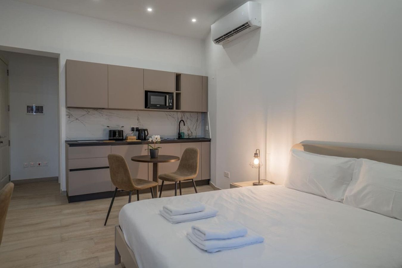 A hotel room with a bed, a bedside lamp, a small table with chairs, a kitchenette with a microwave, and an air conditioning unit.