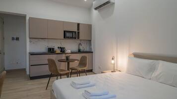 A hotel room with a bed, a bedside lamp, a small table with chairs, a kitchenette with a microwave, and an air conditioning unit.