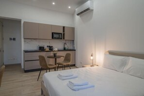 A hotel room with a bed, a bedside lamp, a small table with chairs, a kitchenette with a microwave, and an air conditioning unit.
