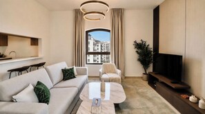 Un salon moderne comprenant un canapé, un fauteuil, une table basse, un téléviseur et une grande fenêtre donnant sur des immeubles.