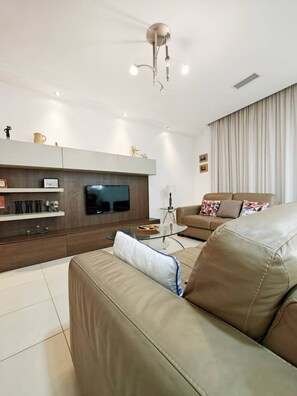 A modern living room with a leather sofa, a flat-screen TV, a glass coffee table, and a ceiling light fixture. - Good Times apartment (Sliema)