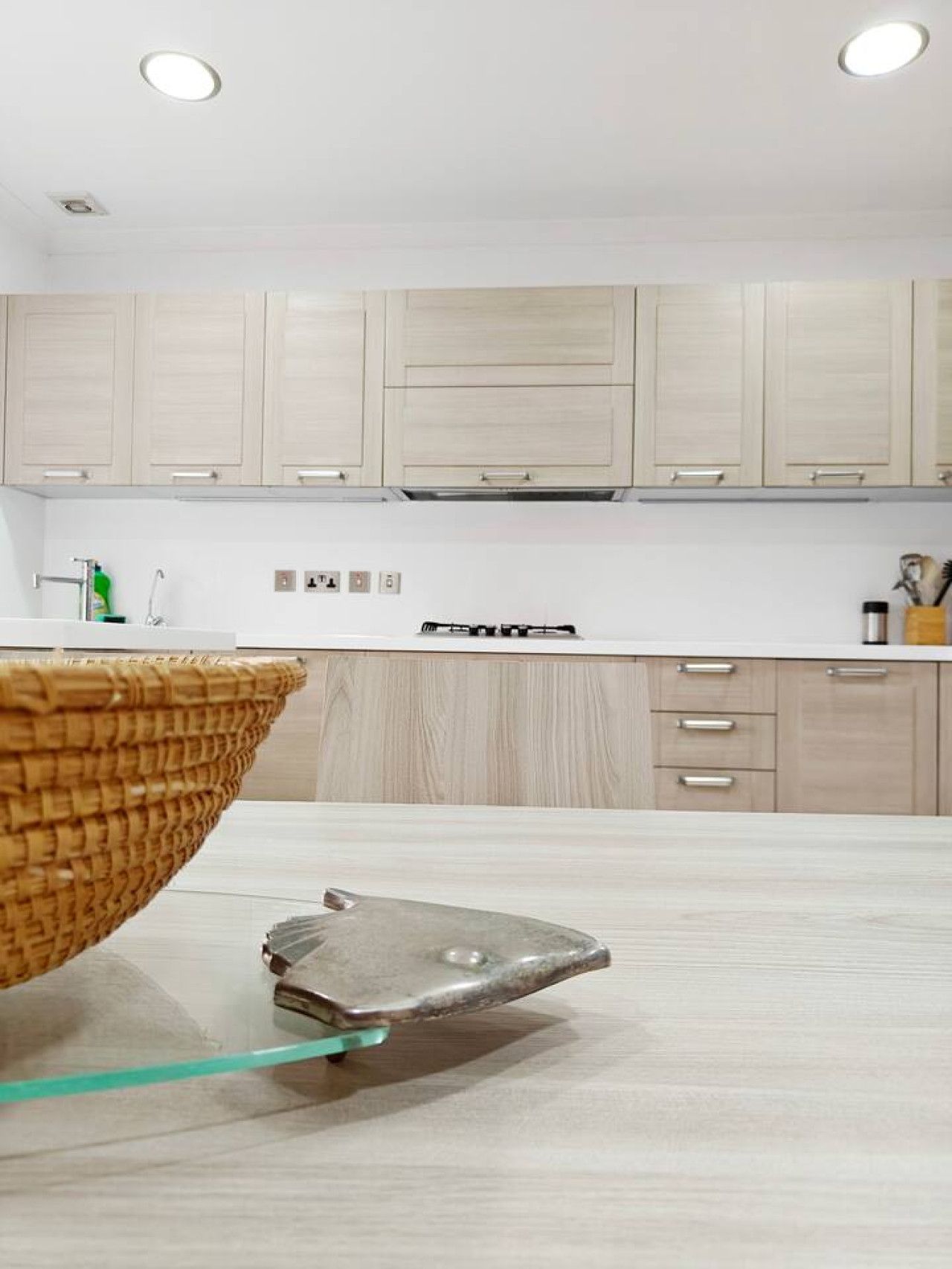 Une cuisine moderne avec des meubles de couleur bois clair, une hotte de cuisine en acier inoxydable et une table en verre surmontée d’un plateau en métal.