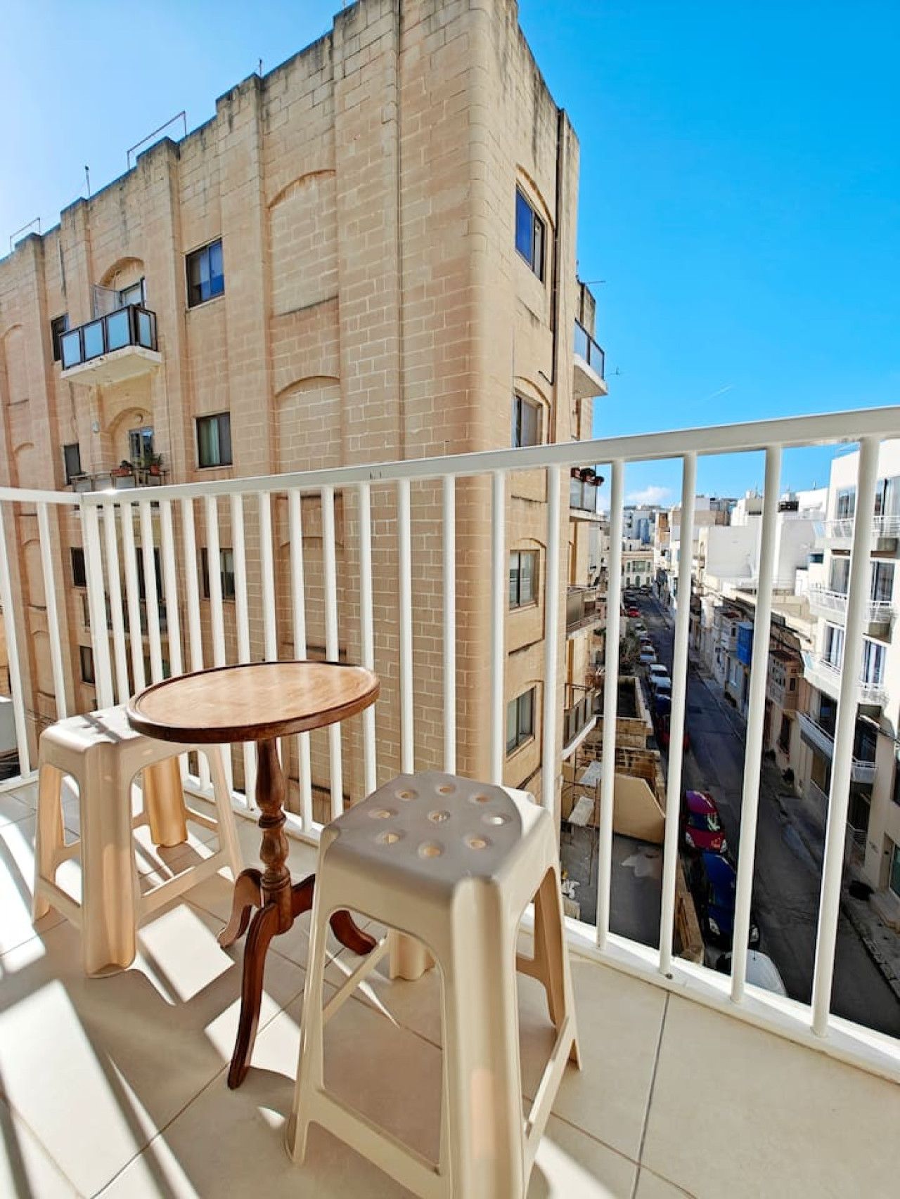 A balcony with a round table and two stools, overlooking a street with buildings.