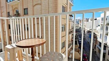 A balcony with a round table and two stools, overlooking a street with buildings.