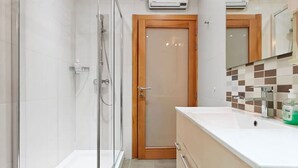 Ein modernes Badezimmer mit einer Duschkabine aus Glas, einer Holztür, einem weißen Waschbecken und einer gefliesten Rückwand.