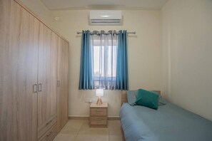 A bedroom with a wooden wardrobe, a bedside table with a lamp, a bed with a blue bedspread, and a window with blue curtains.