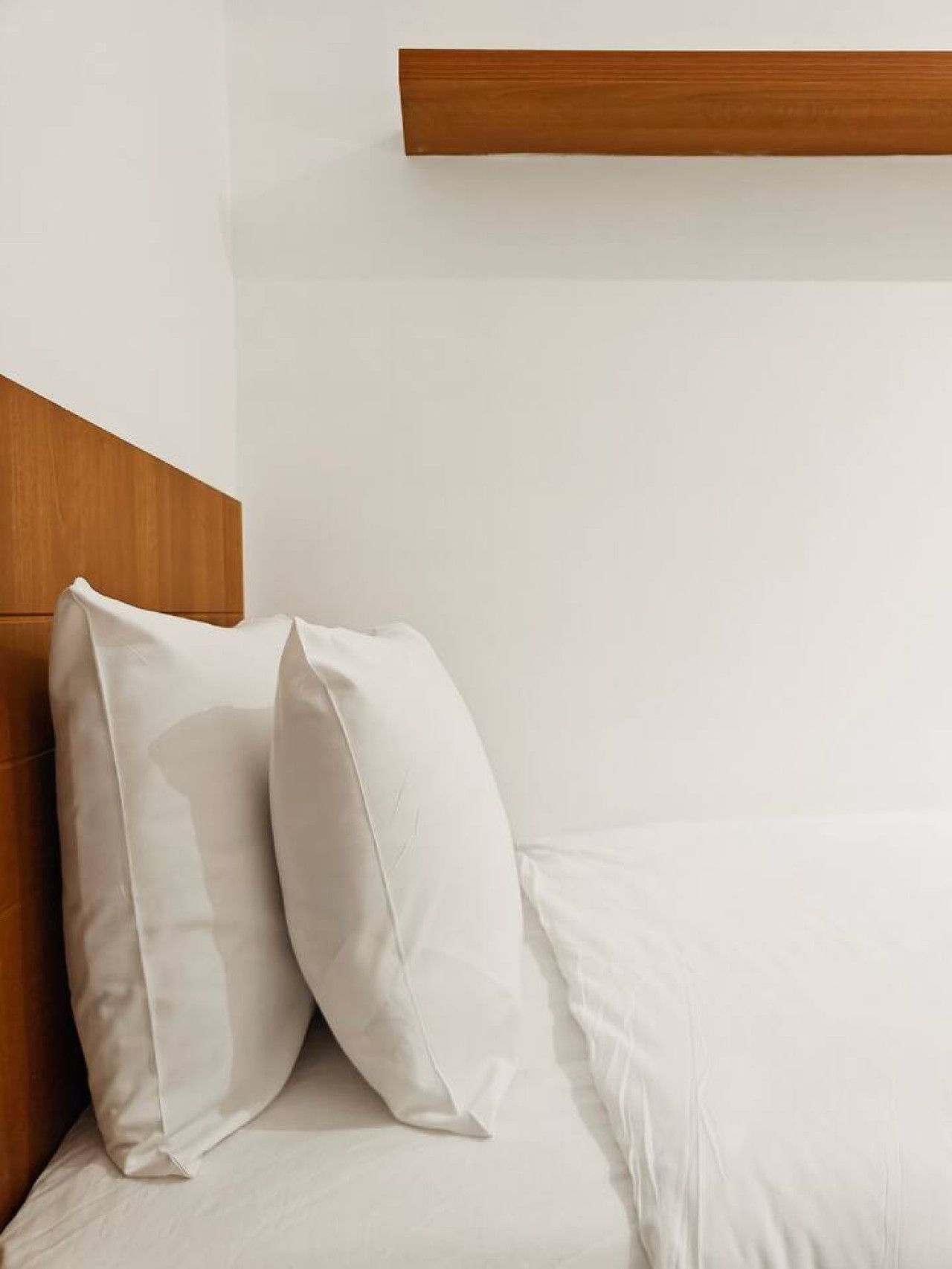 A bed with white linens and two pillows, a wooden headboard, and a wooden shelf above the bed.