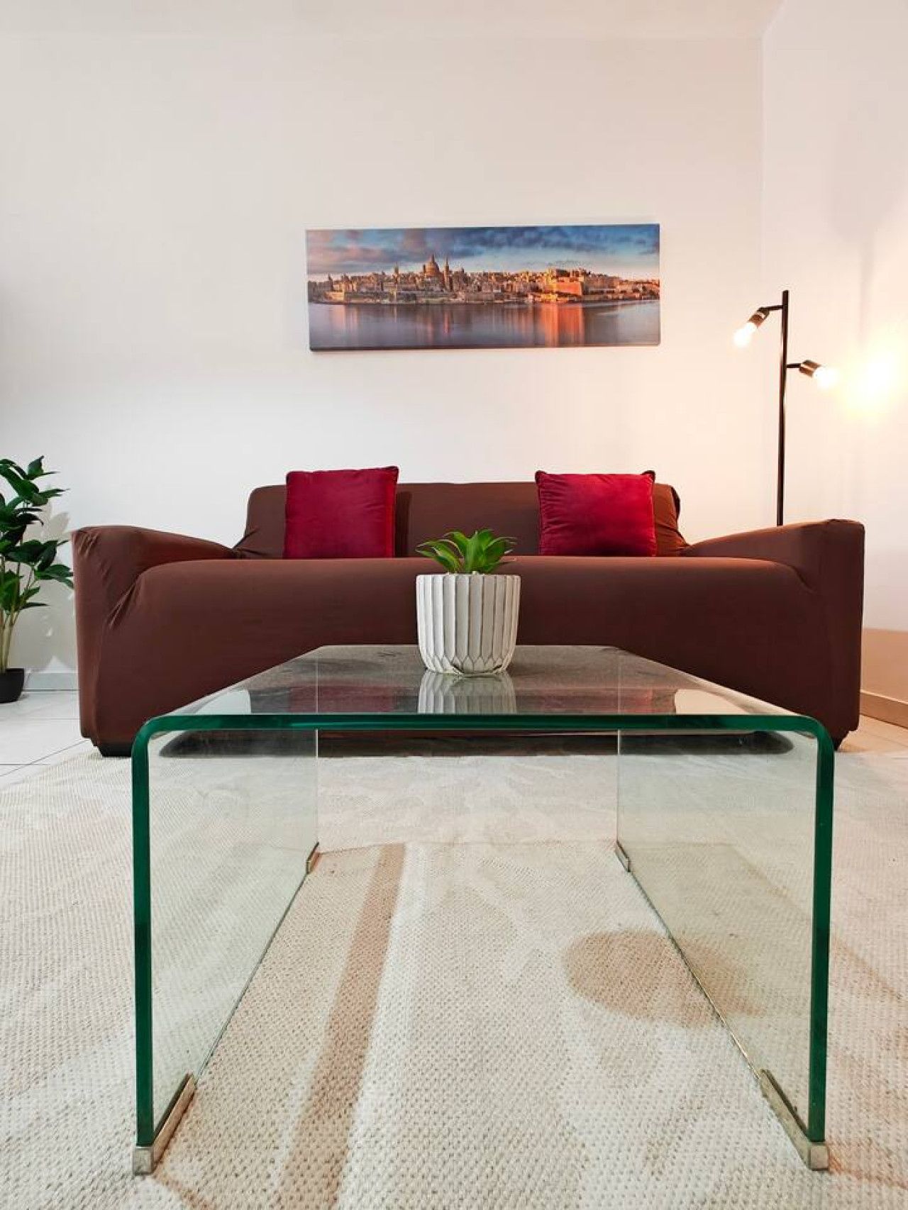 Ein modernes Wohnzimmer mit einem Glastisch, einem braunen Sofa, roten Kissen und einem Stadtbild an der Wand.