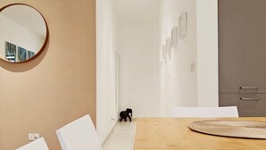 Un interior moderno con una mesa de comedor de madera, sillas blancas, un espejo redondo y un pasillo que conduce a una zona de cocina.