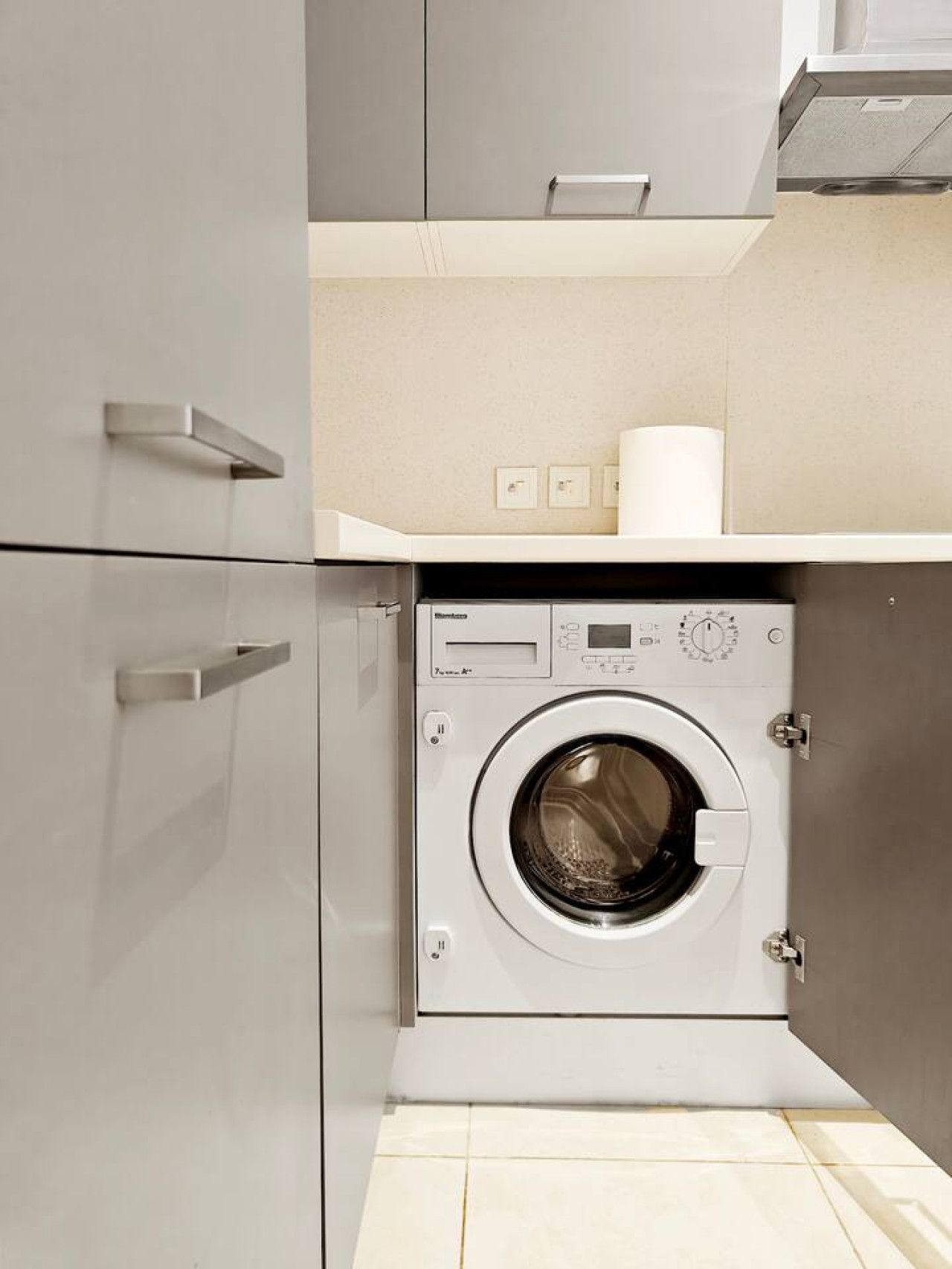 A compact laundry room with a front-loading washing machine, a small shelf with a roll of paper towels, and a wall-mounted light switch.