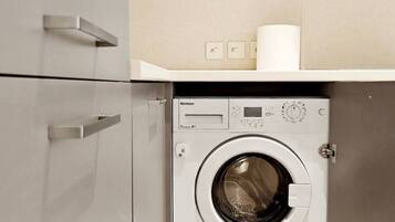 A compact laundry room with a front-loading washing machine, a small shelf with a roll of paper towels, and a wall-mounted light switch.