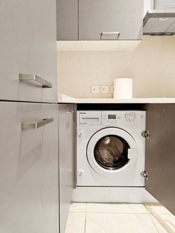 A compact laundry room with a front-loading washing machine, a small shelf with a roll of paper towels, and a wall-mounted light switch.