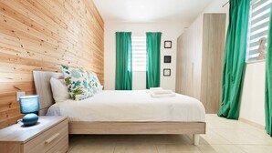 Un dormitorio moderno con cabecera de madera, cama con ropa de cama blanca, cortinas verdes, una mesita de noche con lámpara y un aparador con cajones.