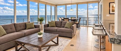 A spacious living area with a large window overlooking the sea, a sofa set, a coffee table, and a dining area with a view.