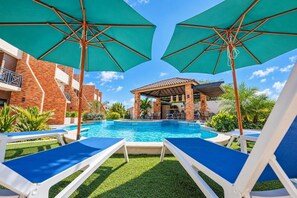 Ein Schwimmbecken mit Liegestühlen und Sonnenschirmen, umgeben von Backsteingebäuden und Grünflächen.