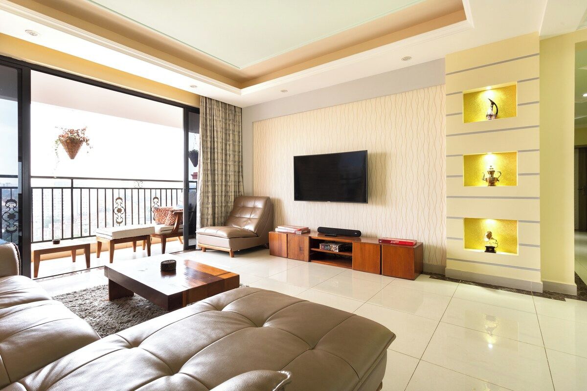 A modern living room with a large sofa, a flat-screen TV, and a balcony with a view.