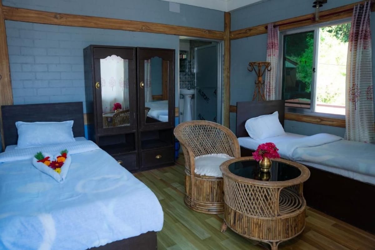 Habitación de hotel con cama, dos sillones de mimbre, una mesita, armario y ventana con cortinas.