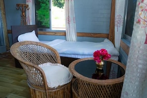 Una habitación con una cama, dos sillones de mimbre, una mesita redonda con un florero y una ventana con cortinas.