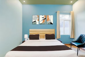 Een slaapkamer met een bed, nachtkastje, lamp, stoel en kunstwerk aan de muur.