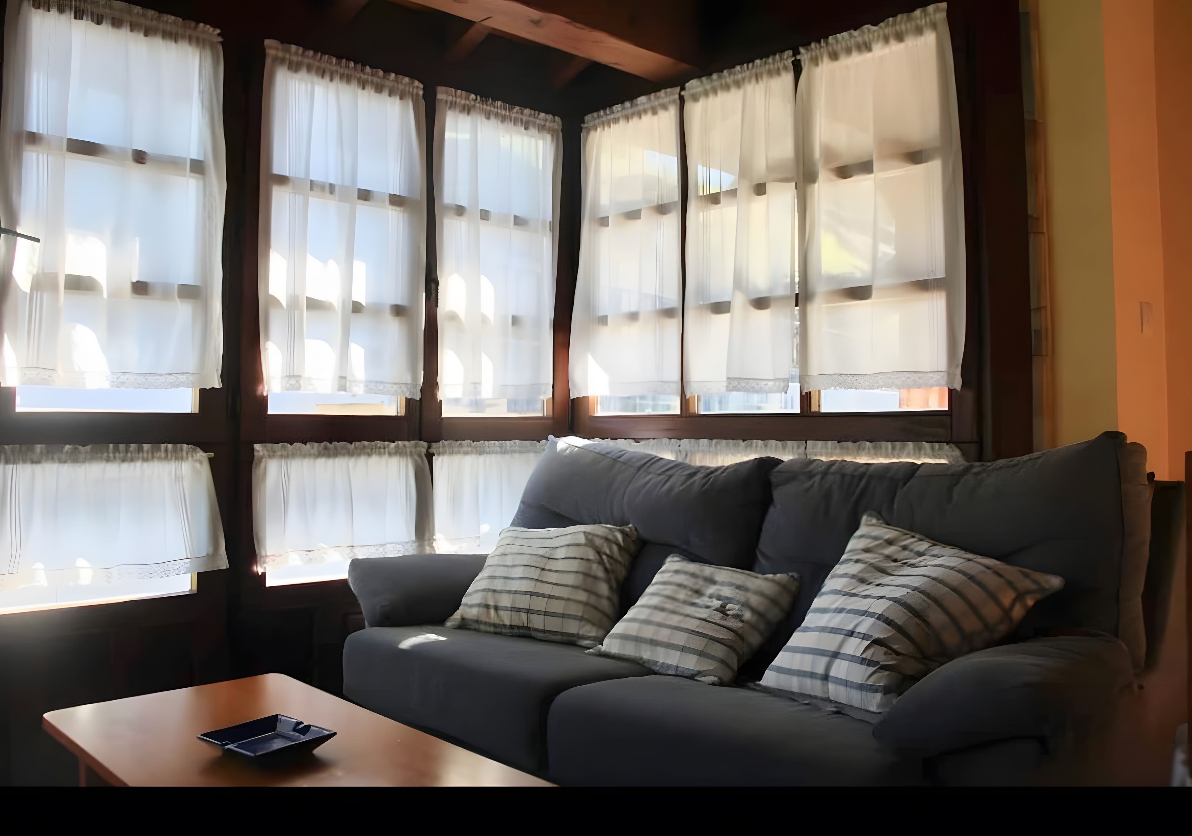 Una habitación con sofá y una mesa, ventanas amplias con cortinas, y vistas al exterior.