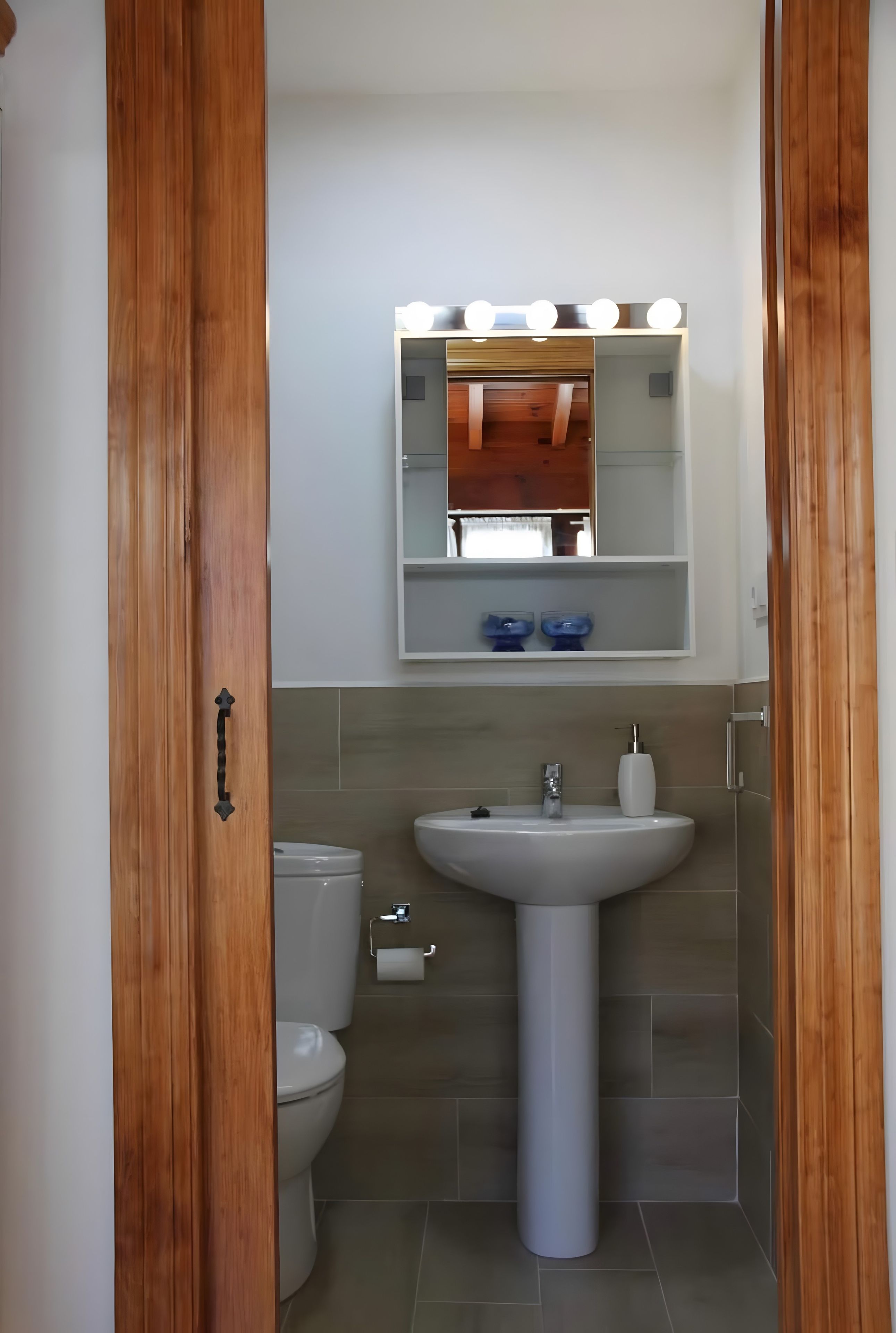 A bathroom with a pedestal sink, a mirror with lights, a toilet, and a wooden door.