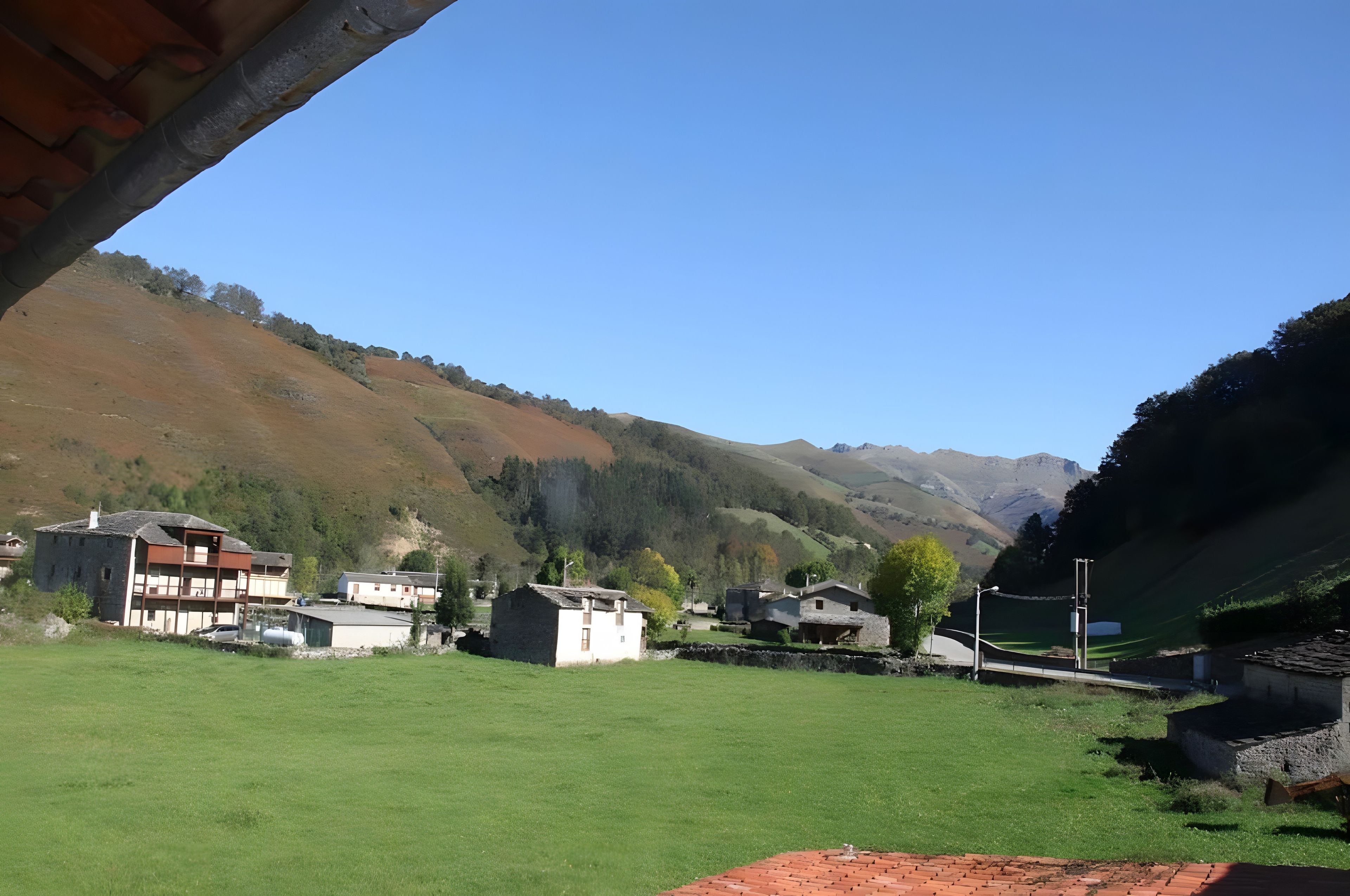 Paesaggio rurale con cielo sereno, un prato erboso e alcuni edifici sullo sfondo di colline.