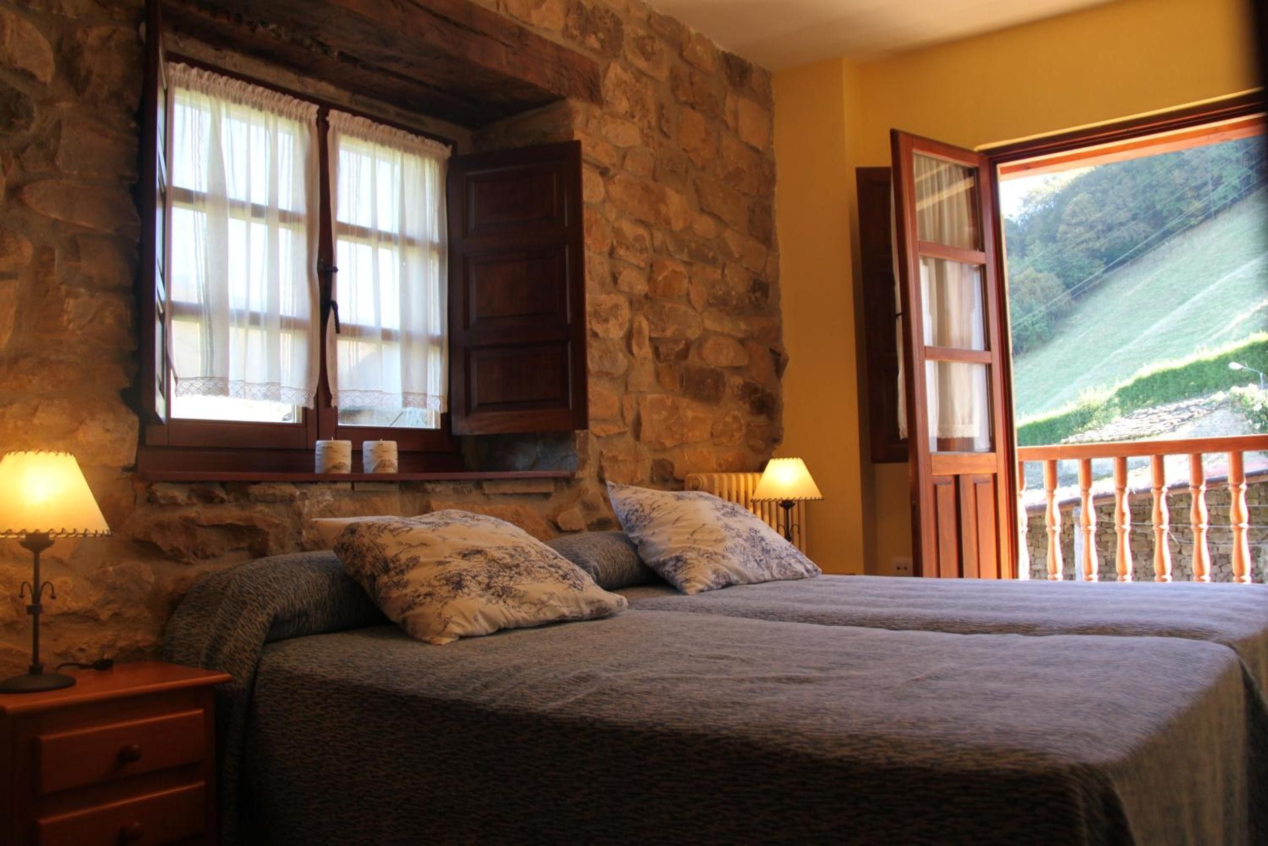 Un dormitorio con pared de piedra, una cama con dos almohadas, una mesita de noche, una ventana con cortinas y vista a un paisaje desde una puerta abierta.