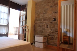 Una habitación con una cama, una silla de madera, una pared de piedra y una mesita con una lámpara.