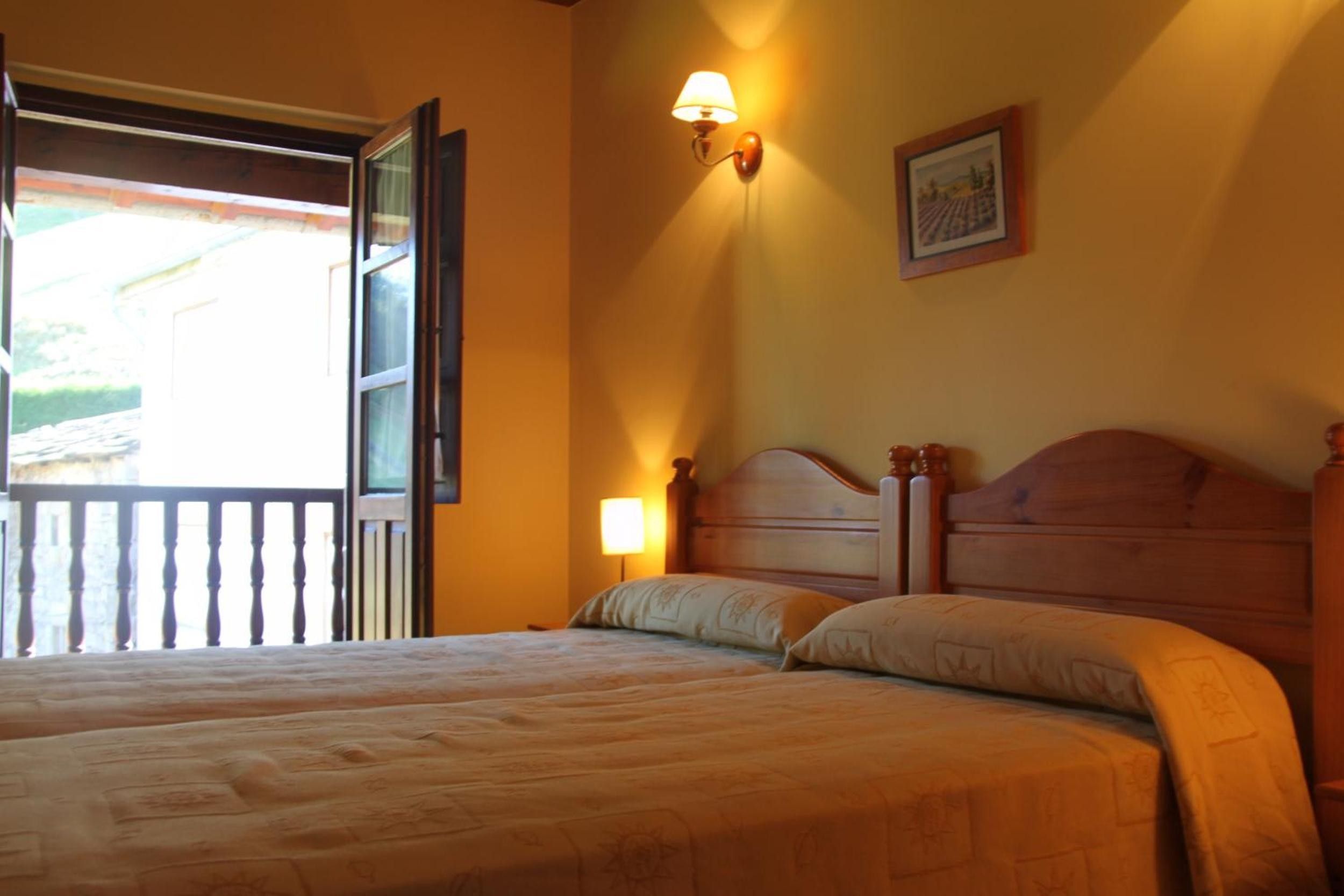Un lit double avec une tête de lit en bois, une table de chevet, une lampe fixée au mur et une fenêtre avec une porte ouverte.
