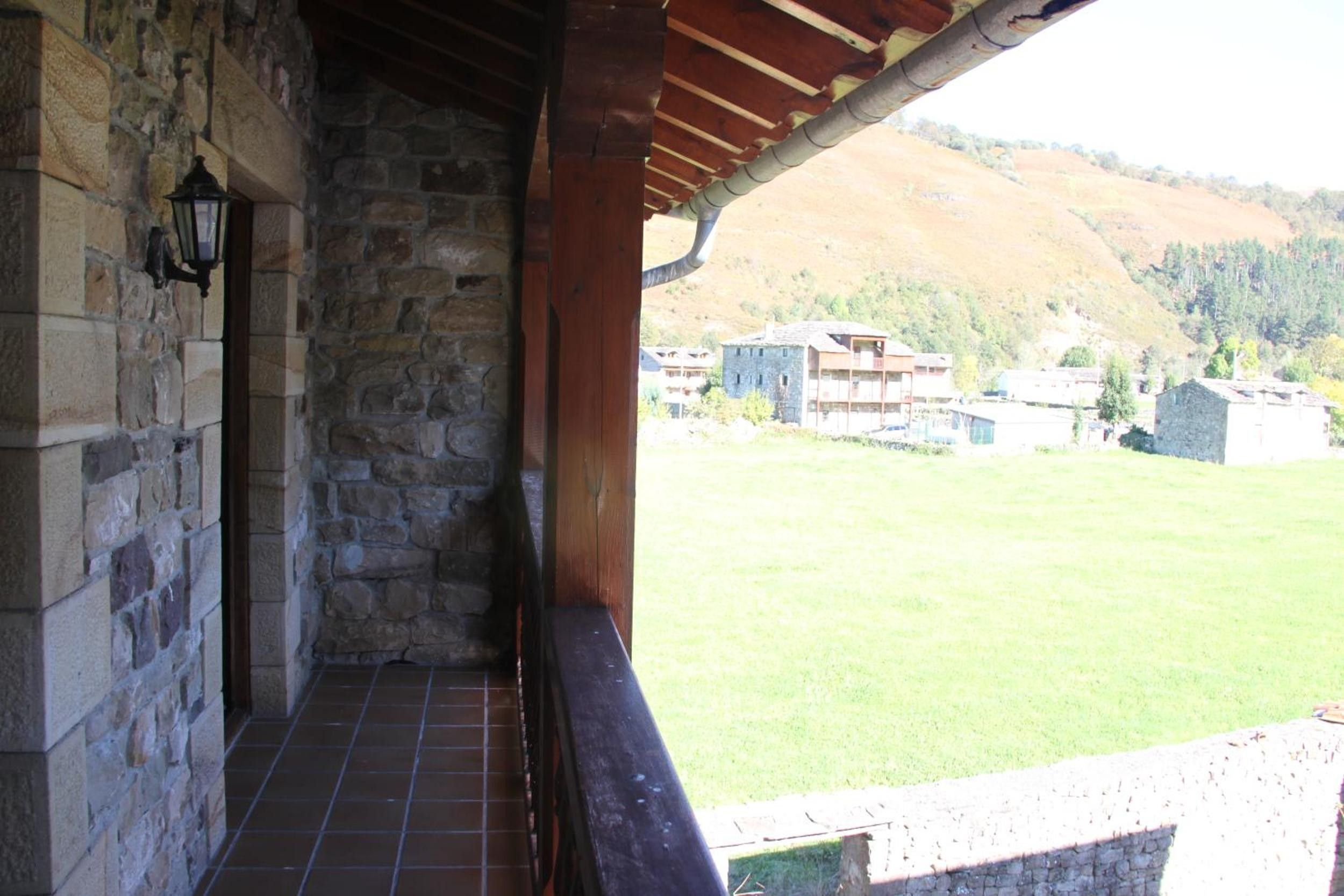 Un edificio de piedra con piso de baldosas y techo de madera. Se aprecia una vista de un campo cubierto de hierba y varios edificios a lo lejos.