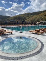 Zona de piscina en un resort con sillones reclinables, un jacuzzi y vistas a una ladera boscosa.