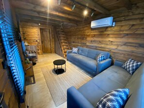 A cozy log cabin interior with a blue sofa, a small round table, and a staircase leading to a loft area. - King Tiny House (Sile)