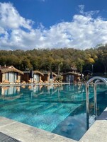 Piscina transparente com espreguiçadeiras e guarda-sóis, cercada por árvores e um edifício com telhado de palha.