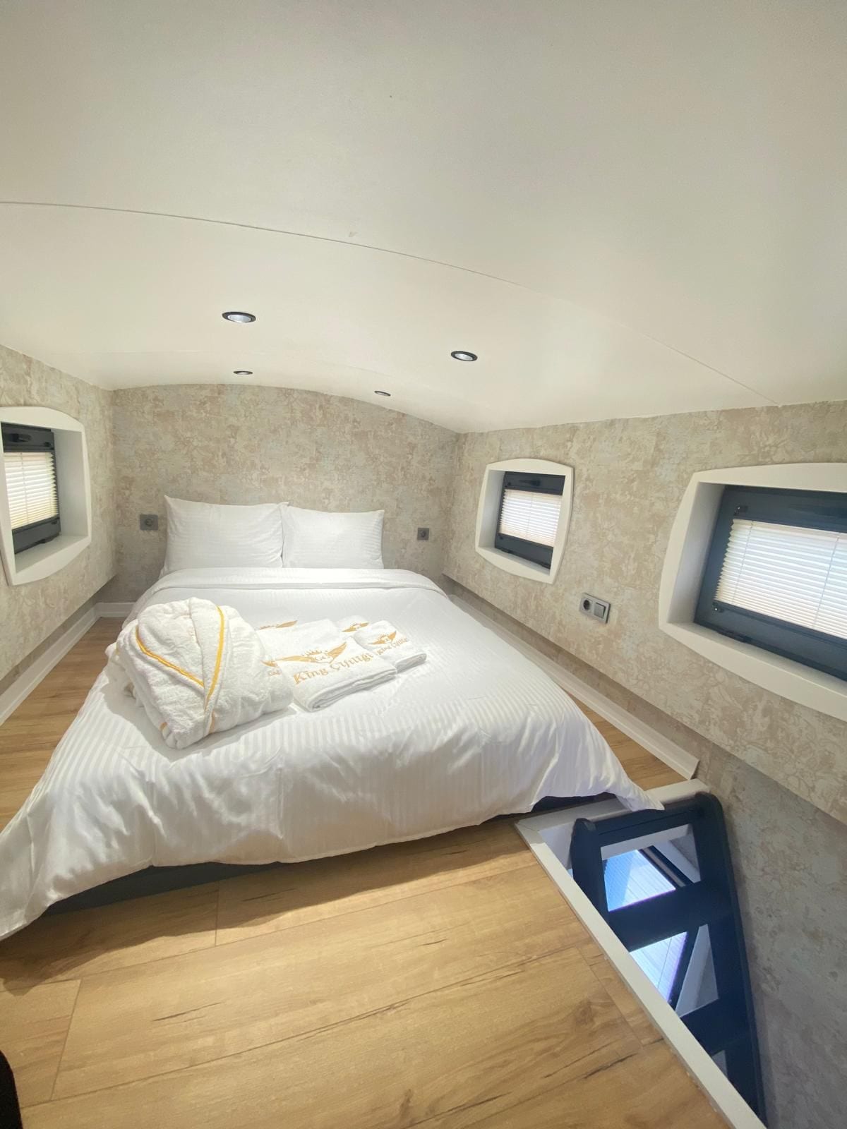 Una habitación de hotel con una cama grande, dos ventanas con persianas, un techo con iluminación empotrada y suelo de madera.