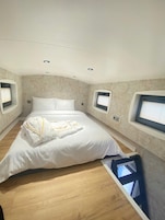 Una habitación de hotel con una cama grande, dos ventanas con persianas, un techo con iluminación empotrada y suelo de madera.