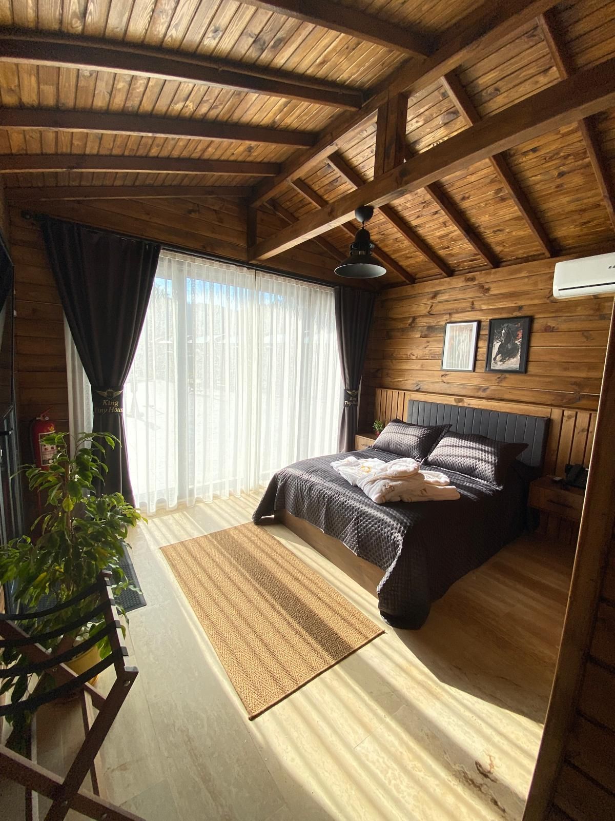 Ein Schlafzimmer mit Holzboden, einem Bett mit dunklem Kopfteil, einem Nachttisch, einer Topfpflanze und einem Fenster mit Vorhängen.