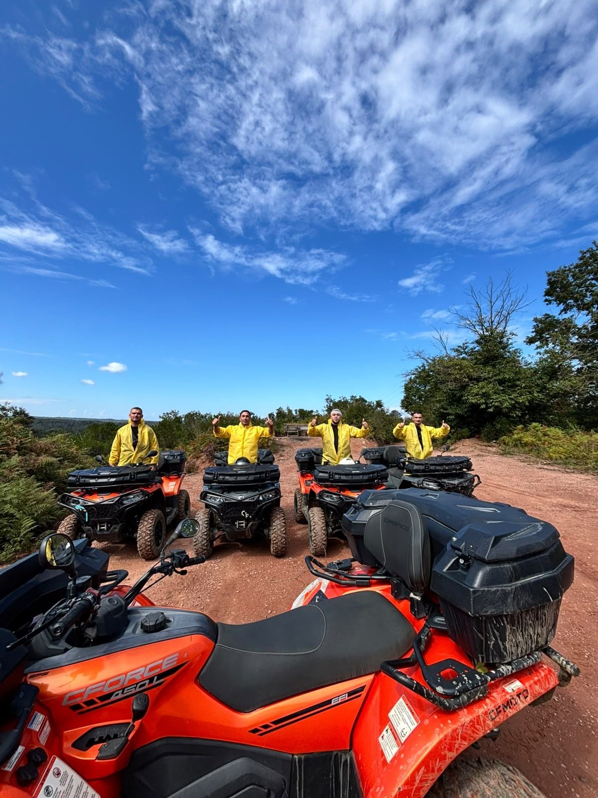 Four people in yellow jackets standing in front of four orange off-road vehicles on a dirt path.