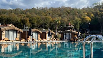 A clear swimming pool with lounge chairs and thatched-roof cabins, surrounded by a forested hillside.