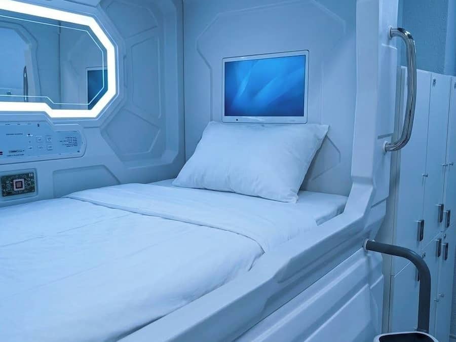 Una habitación futurista con una cama, un panel de control, un monitor y un pequeño taburete.