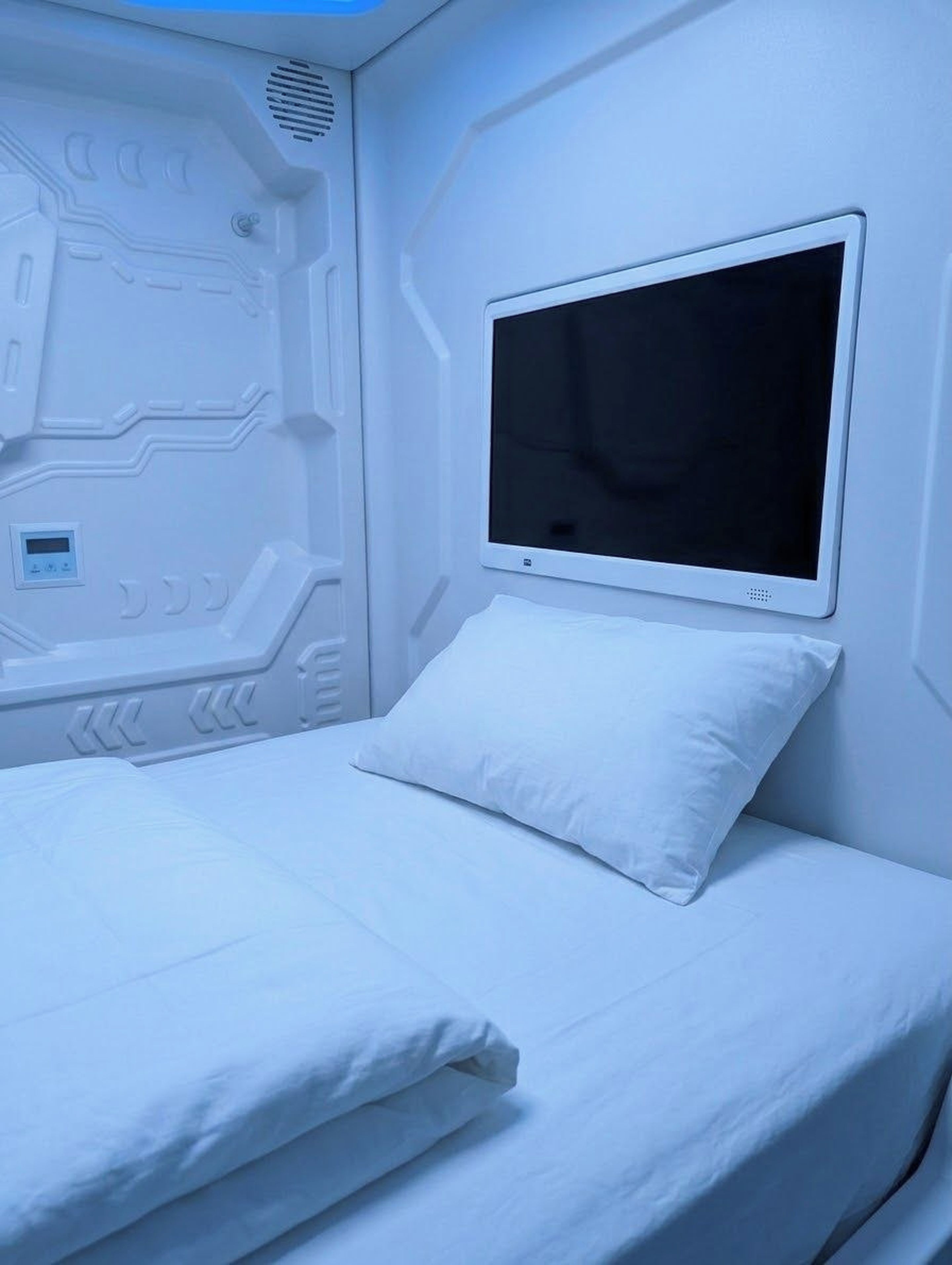 Una cama con colchón y almohada blancos, un televisor de pantalla plana montado en la pared y un panel de control con pantalla digital.