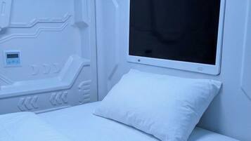 Una cama con colchón y almohada blancos, un televisor de pantalla plana montado en la pared y un panel de control con pantalla digital.