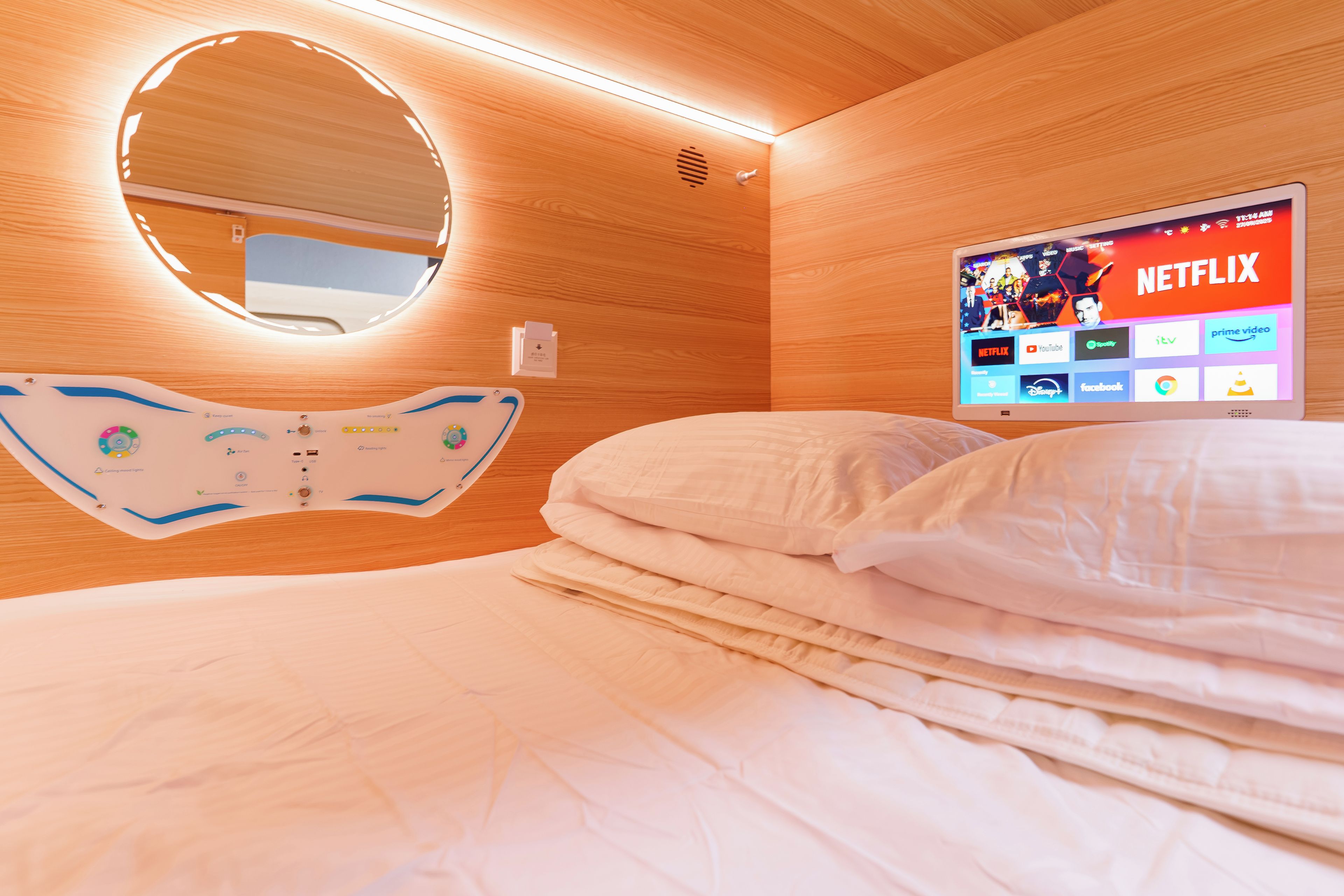 Une chambre d’hôtel comprenant un lit, une table de chevet, un téléviseur fixé au mur affichant l’interface Netflix, et un miroir rond placé au-dessus du lit.