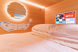 A hotel room with a bed, a bedside table, a wall-mounted TV displaying a Netflix interface, and a circular mirror above the bed.