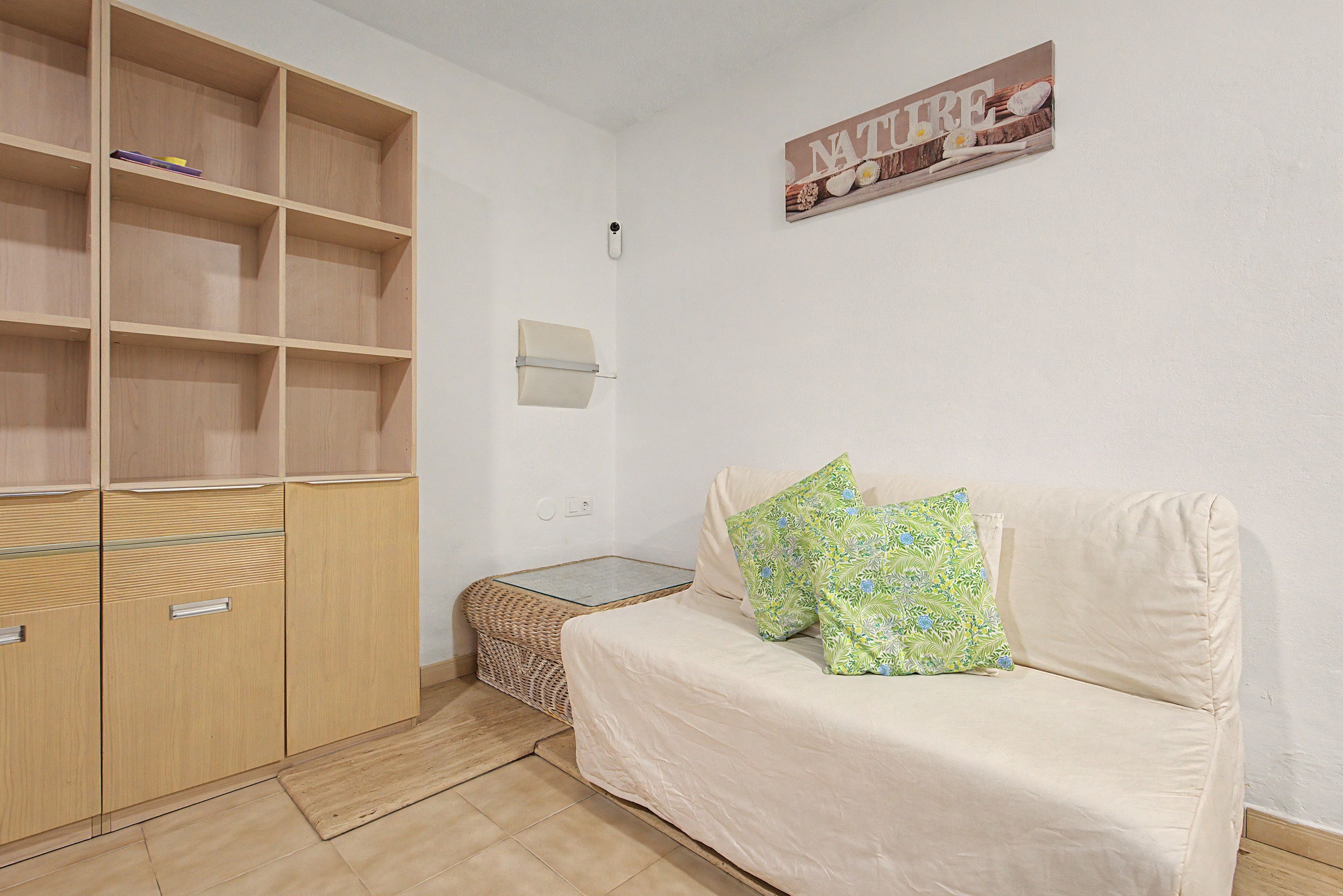 Une pièce avec un canapé beige, deux coussins à motifs, une petite table en osier et un meuble en bois doté d’étagères.