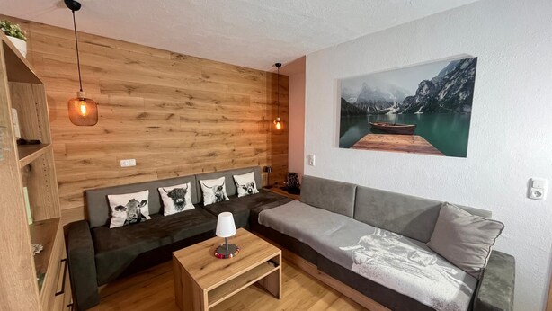 A cozy living room with a wooden wall, a gray sofa, a wooden coffee table, and a mountain landscape painting. - Vrbo Property (Oberreute)